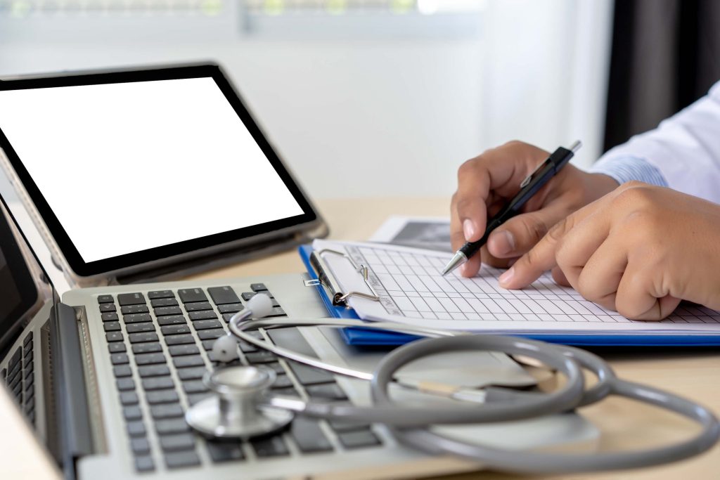 medical professional filling out paperwork using two laptops as reference